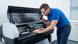 Professional technician in a blue polo shirt and safety glasses inspecting the internal mechanical carriage of a wide-format plotter printer with a flashlight in a clean office setting