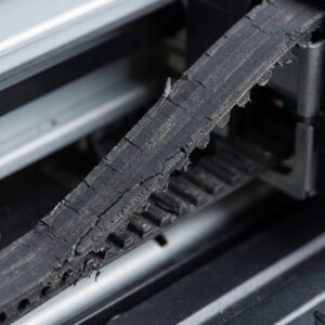 a frayed and worn black rubber carriage belt from a wide-format plotter printer, showing visible cracks and damage to its edges and teeth